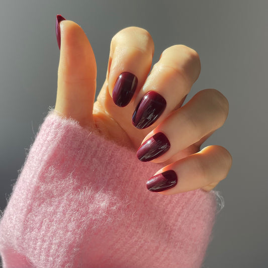 Womans hand wearing sour grape press-on acrylic nails in Candy Claws short oval style. Rich deep plum colour